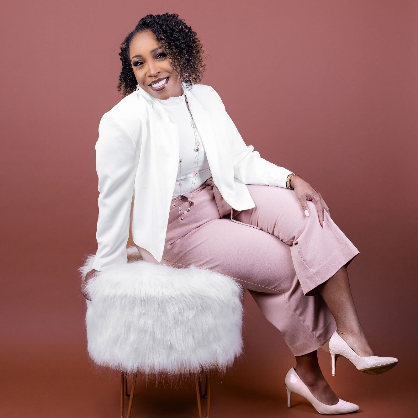 Coaching, Consulting & Retreats for Clarity, Structure and Renewal A woman in a white jacket and pink pants sits confidently on a fluffy stool.